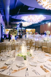 A lavishly decorated banquet hall features multiple round tables adorned with elegant table settings, including white plates, glasses, and beige napkins. The tables are centered with cylindrical glass vases filled with decorative lights and surrounded by greenery. The ambiance is enhanced by large, ornate chandeliers hanging from the ceiling, casting a warm glow across the room. Chairs with a wooden finish are arranged neatly around the tables, and the background features a softly lit atmosphere with blurred figures and decor.