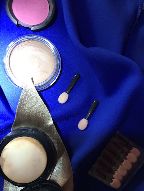 A collection of cosmetic items is arranged on a deep blue fabric. There is a compact powder case, a blush, two small applicator brushes, and a set of lipsticks in a clear case. The items are neatly arranged, with a golden ribbon accentuating the palette.