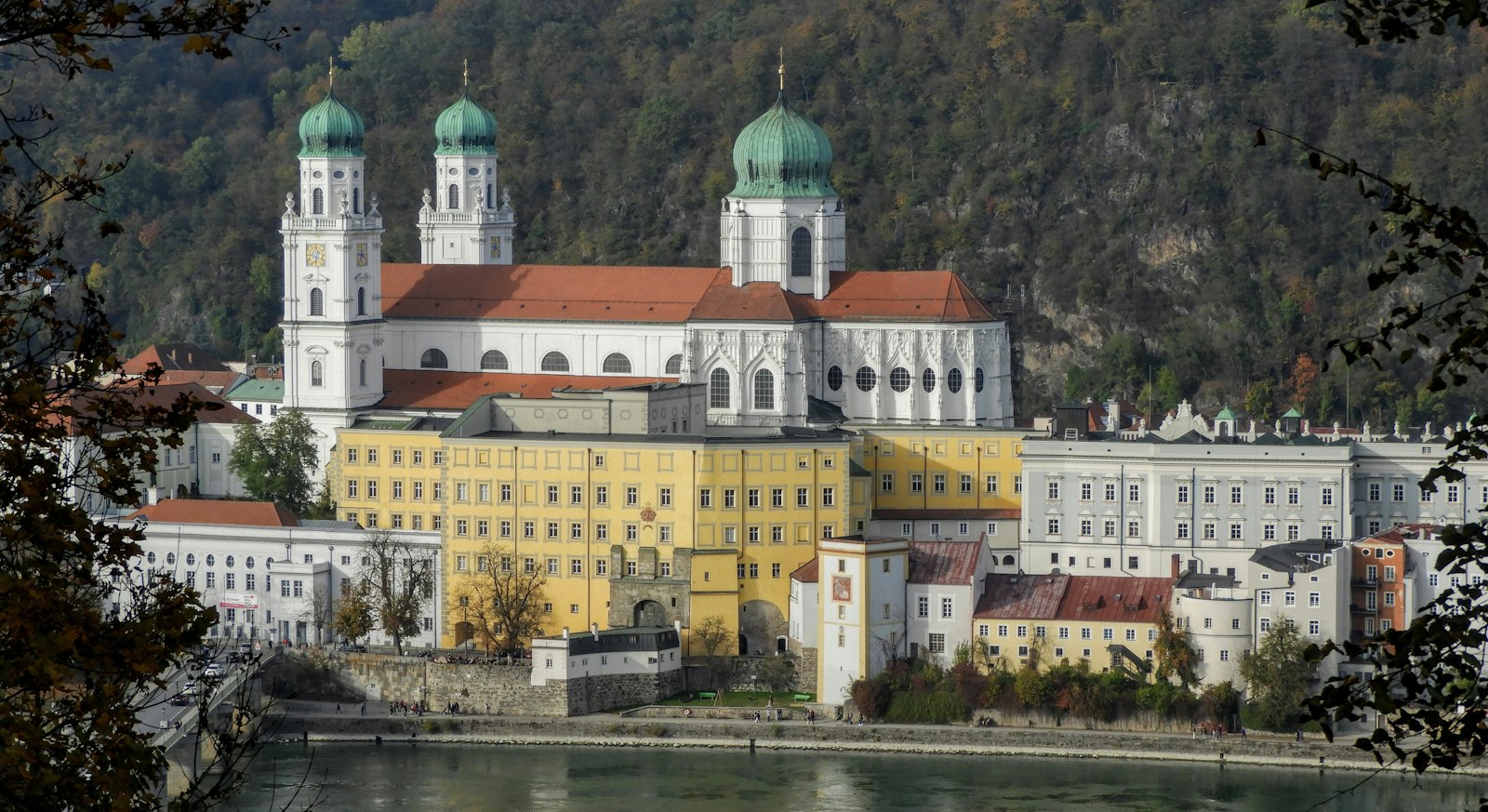 The white towers of St. Stephen's Cathedral rising above pastel Baroque buildings on Passau's riverfront, seen across the Inn through autumn leaves