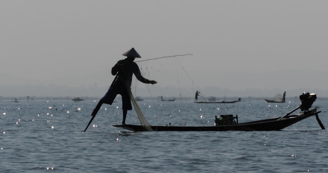 A silhouette of a person fishing on a narrow boat using a unique leg-rowing technique. The water is dotted with sparkling reflections of sunlight, and other fishermen can be vaguely seen in the distance. The atmosphere is calm and serene, with muted colors suggesting early morning or late evening.