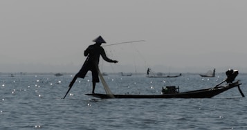 A silhouette of a person fishing on a narrow boat using a unique leg-rowing technique. The water is dotted with sparkling reflections of sunlight, and other fishermen can be vaguely seen in the distance. The atmosphere is calm and serene, with muted colors suggesting early morning or late evening.