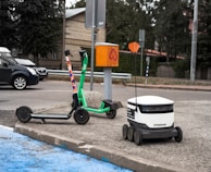 A green e-scooter is parked near a small white delivery robot with 'Starship' branding on it. The scooter and robot are on a sidewalk next to an orange utility box and a street. Behind them is a parked black car and trees lining the street.