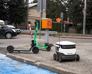 A green e-scooter is parked near a small white delivery robot with 'Starship' branding on it. The scooter and robot are on a sidewalk next to an orange utility box and a street. Behind them is a parked black car and trees lining the street.