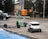 A green e-scooter is parked near a small white delivery robot with 'Starship' branding on it. The scooter and robot are on a sidewalk next to an orange utility box and a street. Behind them is a parked black car and trees lining the street.