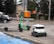 A green e-scooter is parked near a small white delivery robot with 'Starship' branding on it. The scooter and robot are on a sidewalk next to an orange utility box and a street. Behind them is a parked black car and trees lining the street.
