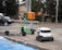 A green e-scooter is parked near a small white delivery robot with 'Starship' branding on it. The scooter and robot are on a sidewalk next to an orange utility box and a street. Behind them is a parked black car and trees lining the street.