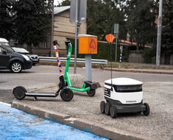 A green e-scooter is parked near a small white delivery robot with 'Starship' branding on it. The scooter and robot are on a sidewalk next to an orange utility box and a street. Behind them is a parked black car and trees lining the street.