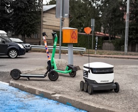 A green e-scooter is parked near a small white delivery robot with 'Starship' branding on it. The scooter and robot are on a sidewalk next to an orange utility box and a street. Behind them is a parked black car and trees lining the street.