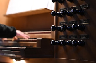 A hand is poised over the keys of a wooden pipe organ, with a detailed view of drawknobs on the side panel. The scene is well-lit, with clear inscriptions on the knobs.
