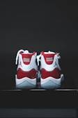 A pair of red and white athletic shoes with a glossy finish, placed on a dark surface. The shoes have white laces and a distinct logo on the heel.
