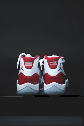 A pair of red and white athletic shoes with a glossy finish, placed on a dark surface. The shoes have white laces and a distinct logo on the heel.