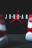The image consists of the word 'JORDAN' with a stylized silhouette of a basketball player in the center. The player is in the iconic jumping pose holding a basketball. The background is dark, and the letters are in white with the silhouette in red. Two blurred objects resembling shoes are in the foreground.