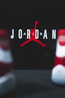 The image consists of the word 'JORDAN' with a stylized silhouette of a basketball player in the center. The player is in the iconic jumping pose holding a basketball. The background is dark, and the letters are in white with the silhouette in red. Two blurred objects resembling shoes are in the foreground.