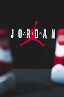 The image consists of the word 'JORDAN' with a stylized silhouette of a basketball player in the center. The player is in the iconic jumping pose holding a basketball. The background is dark, and the letters are in white with the silhouette in red. Two blurred objects resembling shoes are in the foreground.