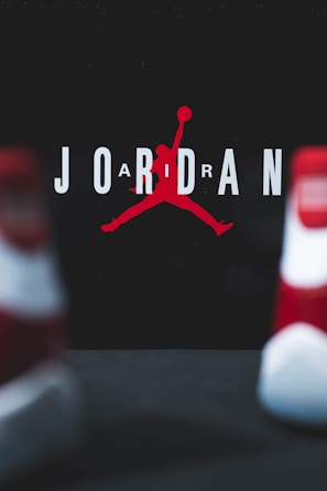The image consists of the word 'JORDAN' with a stylized silhouette of a basketball player in the center. The player is in the iconic jumping pose holding a basketball. The background is dark, and the letters are in white with the silhouette in red. Two blurred objects resembling shoes are in the foreground.