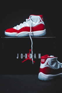 A pair of high-top sneakers with a red, white, and black color scheme is displayed prominently. One shoe is positioned upright on top of a box, while the other lies in front of it. The box is black with the word 'Jordan' and a silhouette of a basketball player printed on it in red and white.