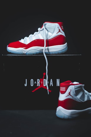 A pair of high-top sneakers with a red, white, and black color scheme is displayed prominently. One shoe is positioned upright on top of a box, while the other lies in front of it. The box is black with the word 'Jordan' and a silhouette of a basketball player printed on it in red and white.
