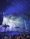A lively night scene at a music festival with a crowd of people gathering under bright, colorful stage lights. Palm trees and fog effects enhance the tropical ambiance, with beams of light creating a dynamic and energetic atmosphere.