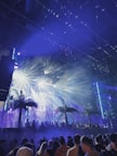 A lively night scene at a music festival with a crowd of people gathering under bright, colorful stage lights. Palm trees and fog effects enhance the tropical ambiance, with beams of light creating a dynamic and energetic atmosphere.