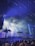 A lively night scene at a music festival with a crowd of people gathering under bright, colorful stage lights. Palm trees and fog effects enhance the tropical ambiance, with beams of light creating a dynamic and energetic atmosphere.