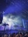 A lively night scene at a music festival with a crowd of people gathering under bright, colorful stage lights. Palm trees and fog effects enhance the tropical ambiance, with beams of light creating a dynamic and energetic atmosphere.