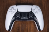 A modern video game controller with a sleek design, featuring white and black colors, with a wooden surface background. The controller includes buttons and joysticks typical for gaming, displaying functionality and style.