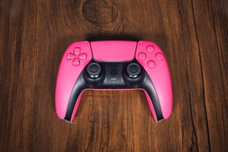 A pink gaming controller with black details, featuring dual analog sticks, directional pad, and action buttons, is positioned centrally on a rich, textured wooden surface.