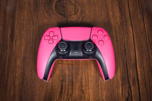 A pink gaming controller with black details, featuring dual analog sticks, directional pad, and action buttons, is positioned centrally on a rich, textured wooden surface.