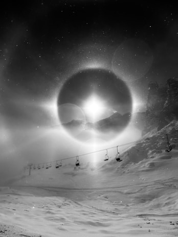 A monochromatic scene depicting a snowy landscape, with a line of empty ski lift chairs suspended in the foreground. A dramatic halo effect encircles the sun, casting ethereal light across the snow-covered ground. Jagged peaks rise in the background, partially obscured by swirling mist.