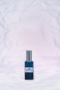 A bottle of perfume with a metallic cap sits on a textured, crumpled white background. The bottle label reads 'ol.factory Aroma Brand' and features a dark, marbled blue design.