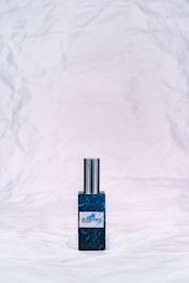 A bottle of perfume with a metallic cap sits on a textured, crumpled white background. The bottle label reads 'ol.factory Aroma Brand' and features a dark, marbled blue design.