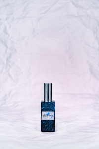 A bottle of perfume with a metallic cap sits on a textured, crumpled white background. The bottle label reads 'ol.factory Aroma Brand' and features a dark, marbled blue design.