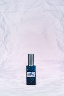 A bottle of perfume with a metallic cap sits on a textured, crumpled white background. The bottle label reads 'ol.factory Aroma Brand' and features a dark, marbled blue design.