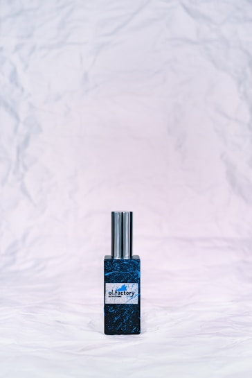 A bottle of perfume with a metallic cap sits on a textured, crumpled white background. The bottle label reads 'ol.factory Aroma Brand' and features a dark, marbled blue design.