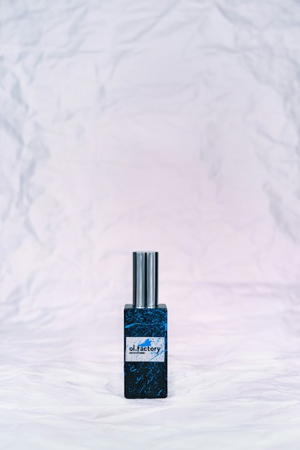 A bottle of perfume with a metallic cap sits on a textured, crumpled white background. The bottle label reads 'ol.factory Aroma Brand' and features a dark, marbled blue design.