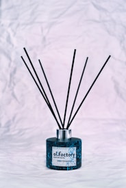 A black and blue decorative fragrance diffuser sits on a crumpled white textured surface. The bottle is medium-sized with thin black reeds extending upwards.