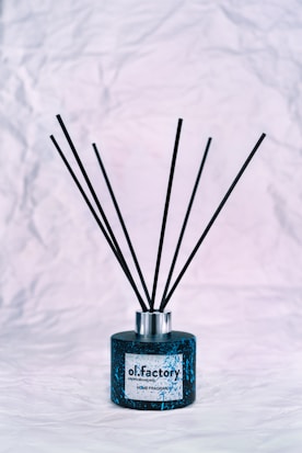 A black and blue decorative fragrance diffuser sits on a crumpled white textured surface. The bottle is medium-sized with thin black reeds extending upwards.
