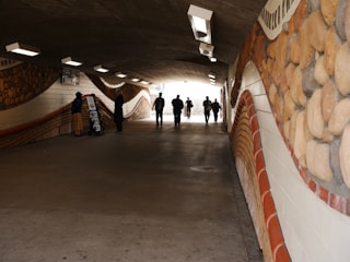 A dimly lit underpass with artistic stone-like murals on the walls. Several people walk or stand along the pathway, and two figures are engaged in conversation near a display stand.