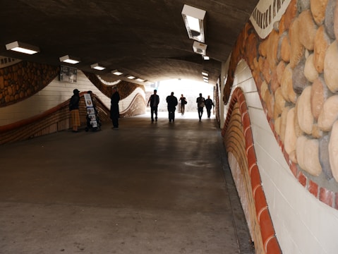 A dimly lit underpass with artistic stone-like murals on the walls. Several people walk or stand along the pathway, and two figures are engaged in conversation near a display stand.