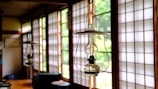 An interior with traditional Japanese sliding doors made of wood and frosted glass or paper, letting in ample natural light. Hanging close to the camera is a glass kerosene lamp, adding a rustic touch to the serene environment. The room has wooden floors and simple furniture, reflecting a minimalist aesthetic.