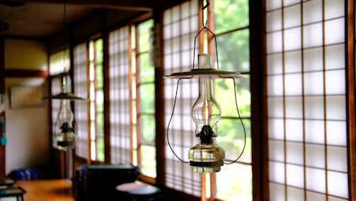 An interior with traditional Japanese sliding doors made of wood and frosted glass or paper, letting in ample natural light. Hanging close to the camera is a glass kerosene lamp, adding a rustic touch to the serene environment. The room has wooden floors and simple furniture, reflecting a minimalist aesthetic.