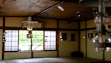 An interior of a traditional room with wooden elements and multiple hanging lanterns. Shoji screens with square patterns partially open to reveal greenery outside. The room has a serene and rustic atmosphere, filled with natural light filtering through the windows.