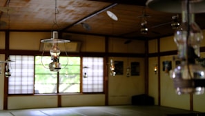 An interior of a traditional room with wooden elements and multiple hanging lanterns. Shoji screens with square patterns partially open to reveal greenery outside. The room has a serene and rustic atmosphere, filled with natural light filtering through the windows.