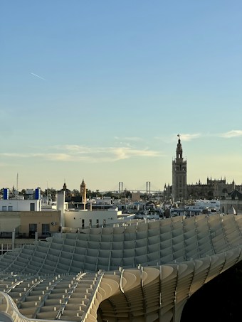 The image depicts a cityscape with a unique architectural structure featuring curved, lattice-like surfaces in the foreground. In the background, there are historic-looking buildings including a tall bell tower and a distant bridge. The sky is clear with a subtle trail of an airplane visible.