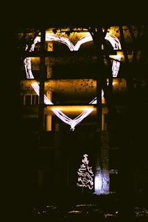 A building façade illuminated with a large outline of a heart in neon lights and a lit Christmas tree below. The scene is enveloped in darkness, with silhouettes of tree branches partially obstructing the view.