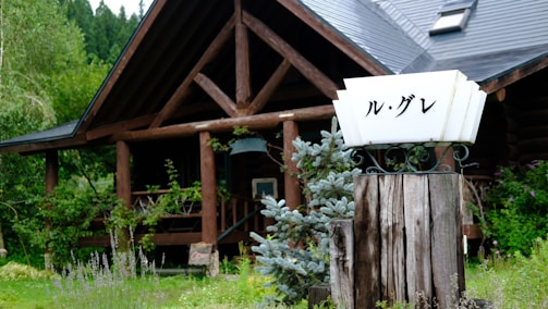 A rustic wooden cabin surrounded by lush green vegetation with a wooden signpost in the foreground displaying Japanese characters. The cabin has a slanted roof and a porch with railing, partially hidden by trees and plants.