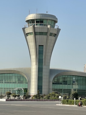 A tall, modern control tower with a cylindrical base standing in front of a building that features large curved windows. Surrounding the building are some green shrubs and trees. The sky overhead is clear and blue.