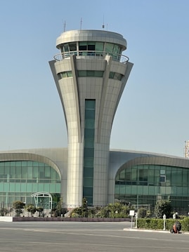 A tall, modern control tower with a cylindrical base standing in front of a building that features large curved windows. Surrounding the building are some green shrubs and trees. The sky overhead is clear and blue.