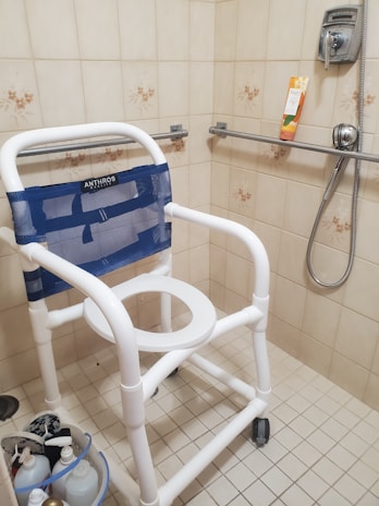 A bathroom setting with a shower chair featuring a white frame and a blue mesh backrest. The chair is designed for accessibility and has wheels for mobility. The surrounding shower area has beige tiled walls with floral accents. A handheld showerhead is mounted on the wall, along with a safety grab bar. Several toiletry bottles are placed in the corner on a shower caddy.