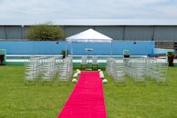 A wedding ceremony setup featuring a white canopy tent at the center with transparent chairs arranged in rows on a green lawn. A red carpet aisle is flanked by white flower arrangements, leading to the canopy. The background includes a blue painted wall, some industrial buildings, and potted plants.